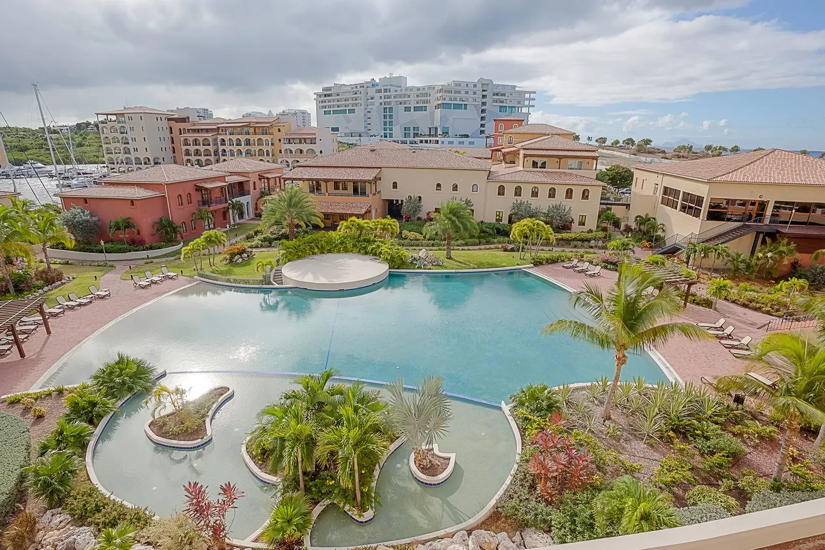 The large resort swimming pool features decorative island planters and sun deck, surrounded by Mediterranean-style buildings and tropical landscaping with harbor views.