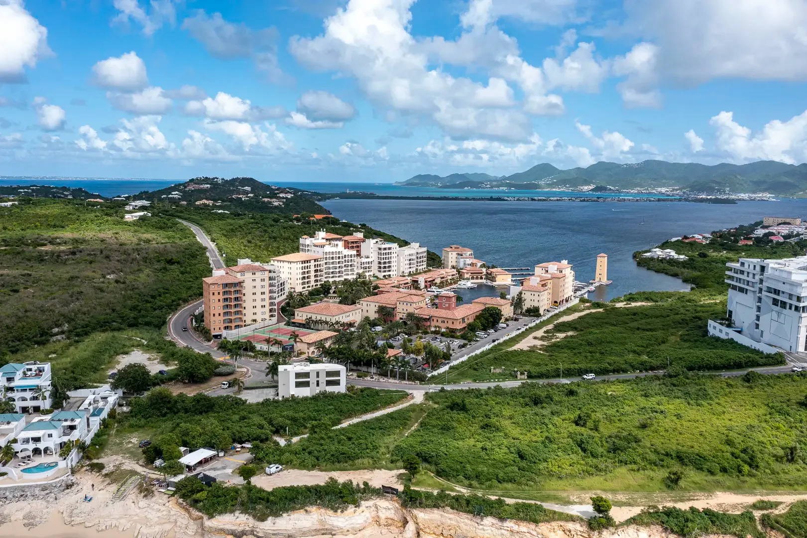 An elevated wide-angle shot captures the resort complex, marina, tennis courts, lush green hills, blue water, and distant mountains.