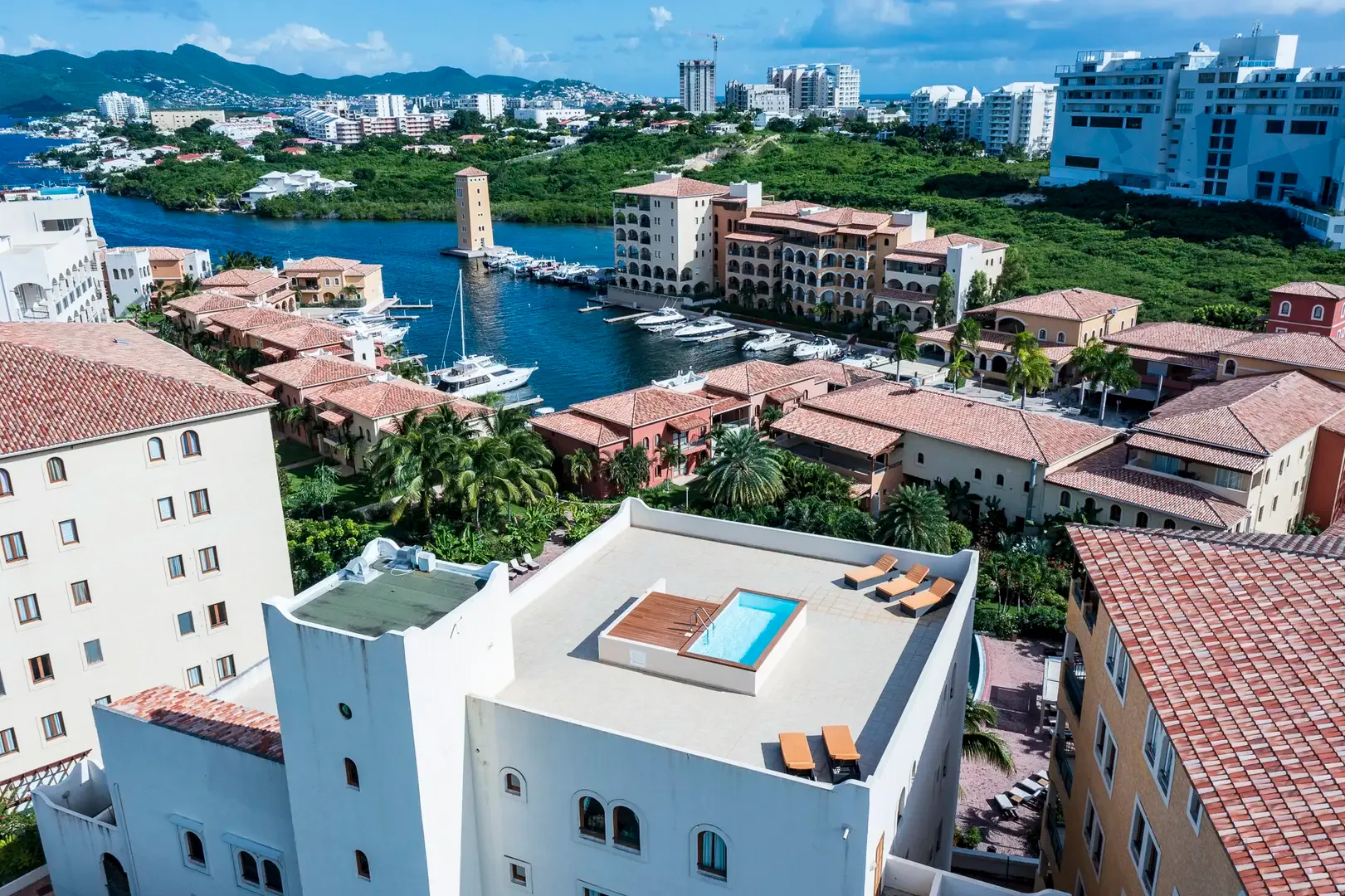 An aerial view reveals your private rooftop pool with lounge chairs, the Mediterranean-style resort complex, marina with yachts, and lush palm trees.