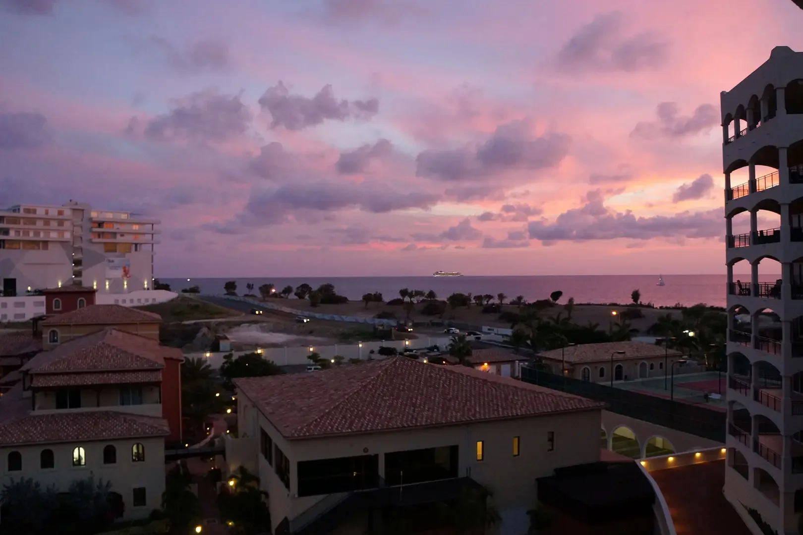 A stunning sunset overlooks the resort grounds and ocean, with vibrant pink and purple clouds, ship on horizon, and ambient building lights.