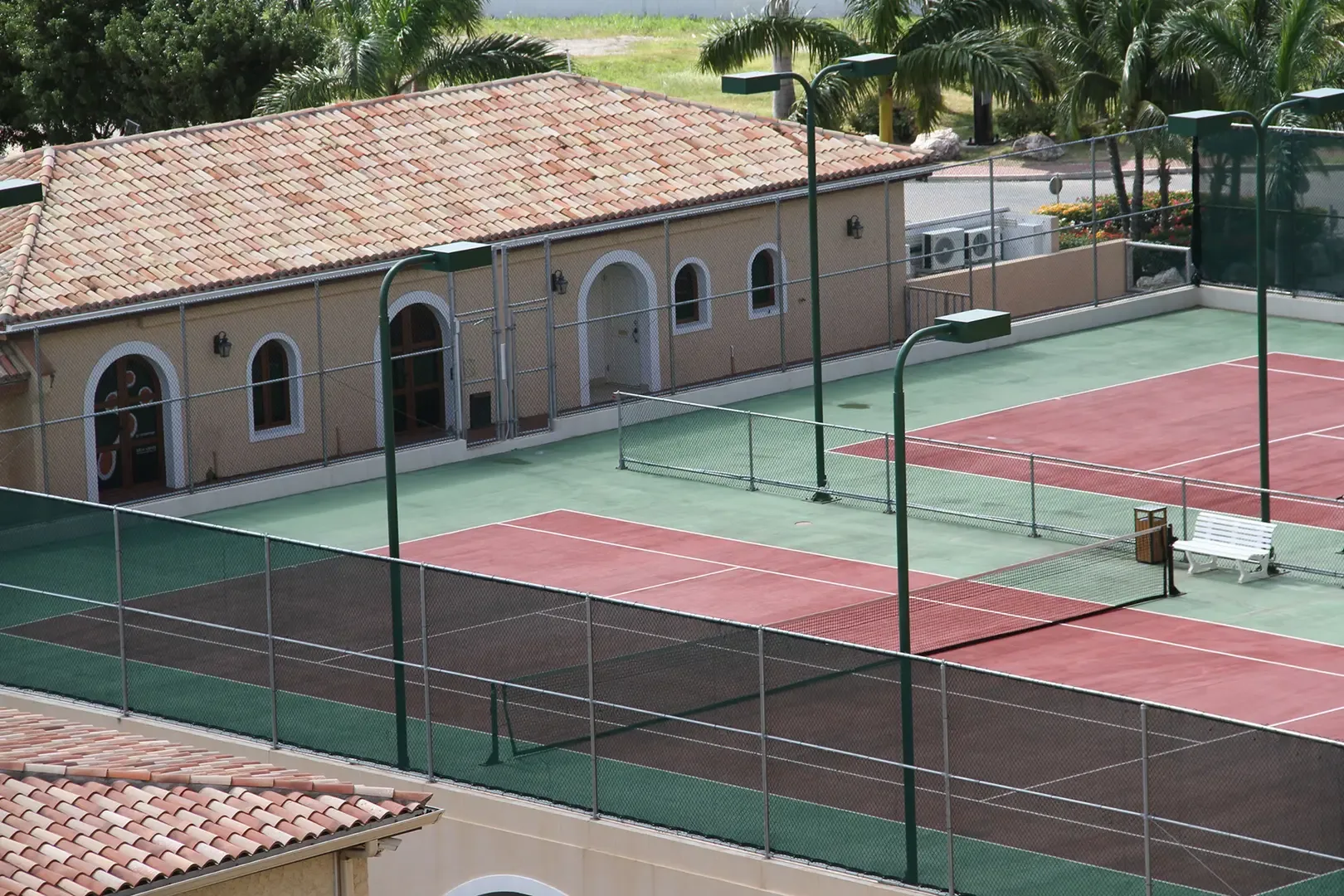 Two outdoor tennis courts with green and red surfaces, tall green light poles, fencing, white bench, and adjacent building with terracotta tile roof.