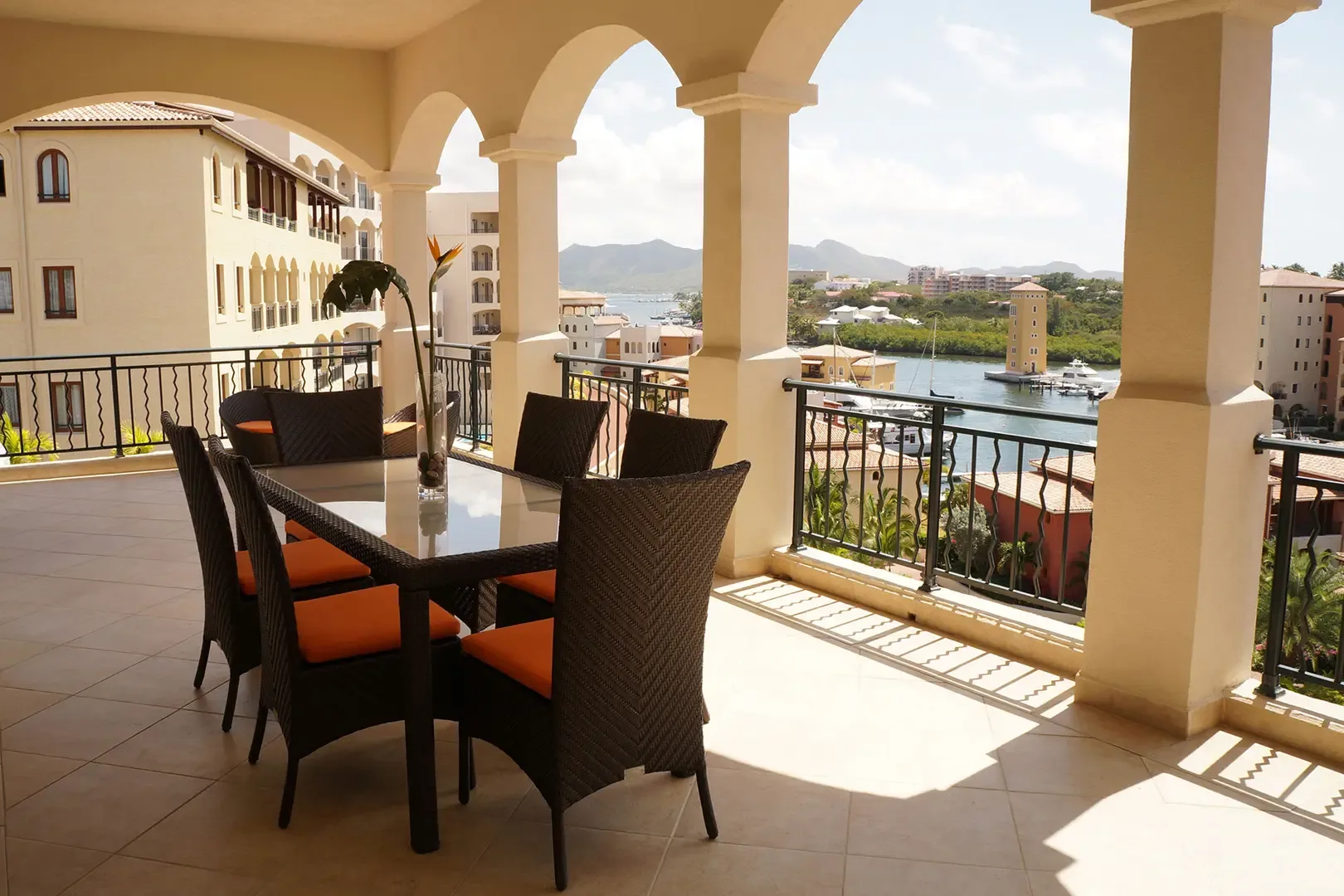 The spacious outdoor terrace features an arched balcony with a dining table and woven chairs with orange cushions, overlooking the marina and tropical landscape.