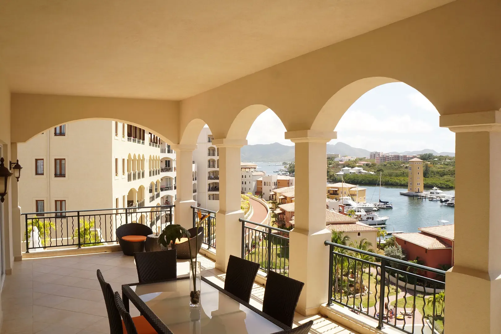 A spacious terrace with dining table, chairs, and lounge area offers elegant arched openings and stunning views of the marina, boats, and distant mountains.