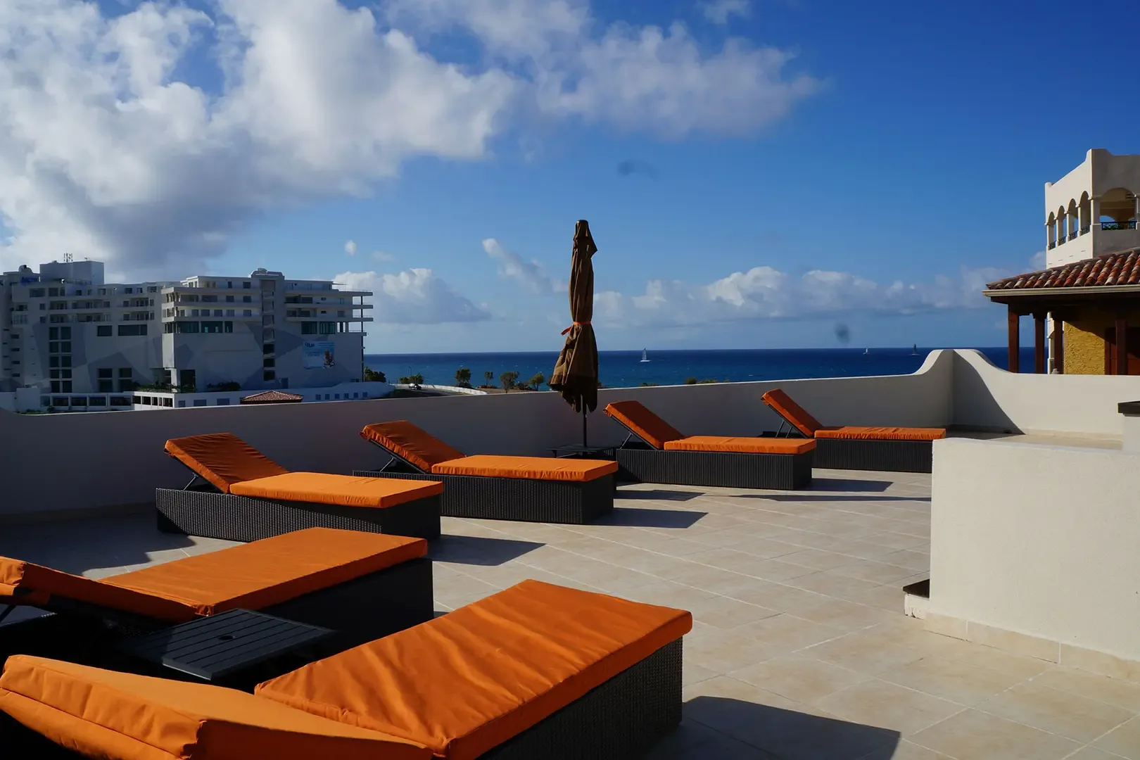 Modern lounge chairs with bright orange cushions line the rooftop terrace, offering sweeping views of the ocean and nearby resort buildings.