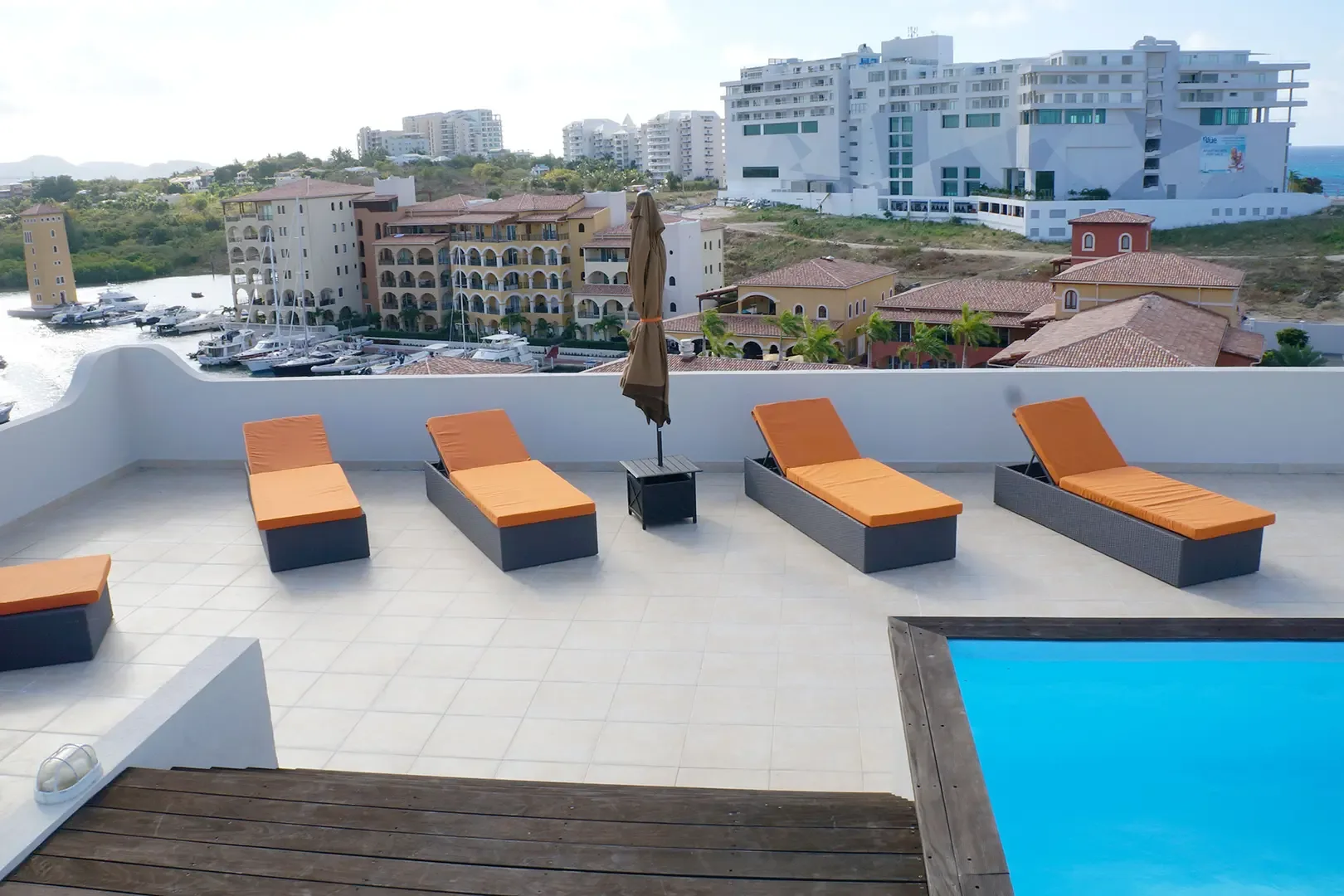 Four orange lounge chairs and a patio umbrella sit beside the infinity pool corner, with harbor views and resort architecture in the background.