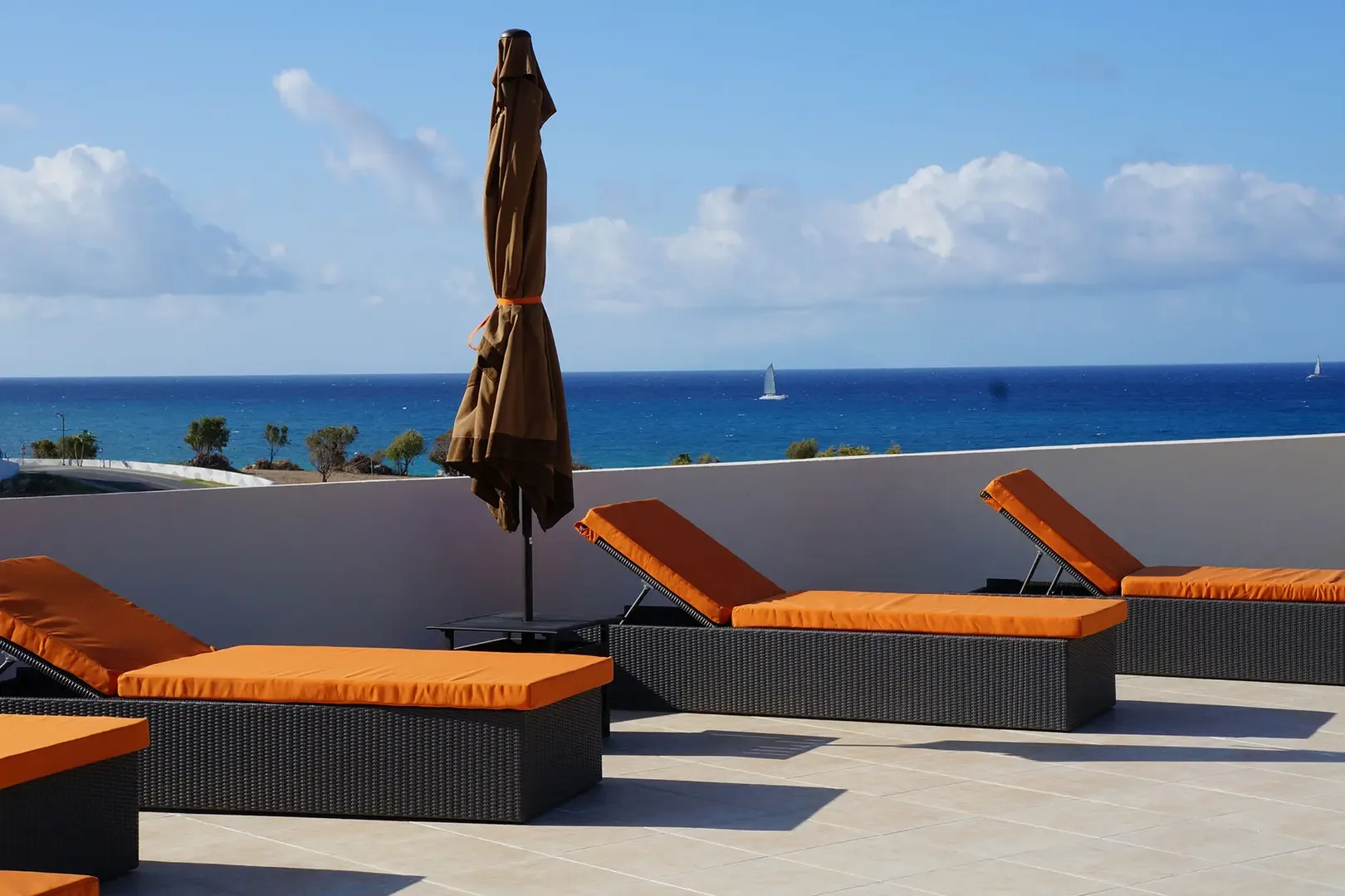 Sun terrace with orange lounge chairs and umbrella overlooking the ocean with sailboat on the horizon.