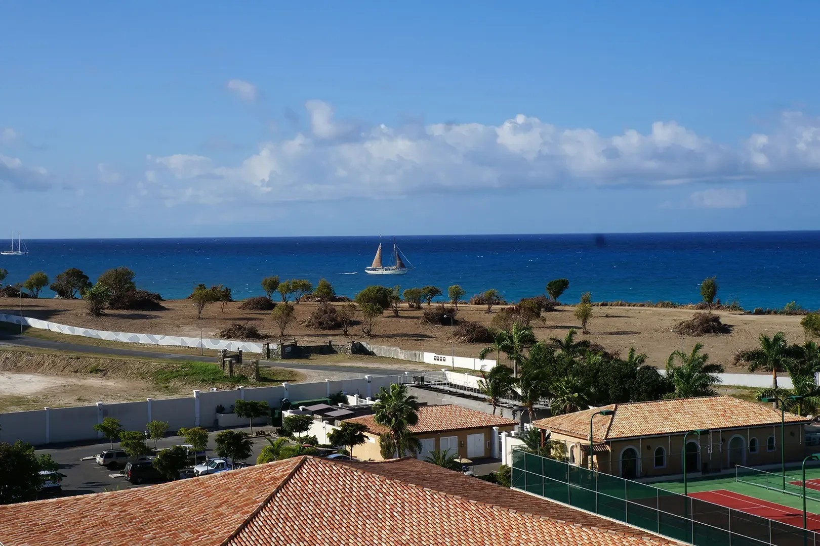Elevated view overlooking coastal development with tiled roofs, tennis court, parking area, and large sailboat on blue ocean in the distance.