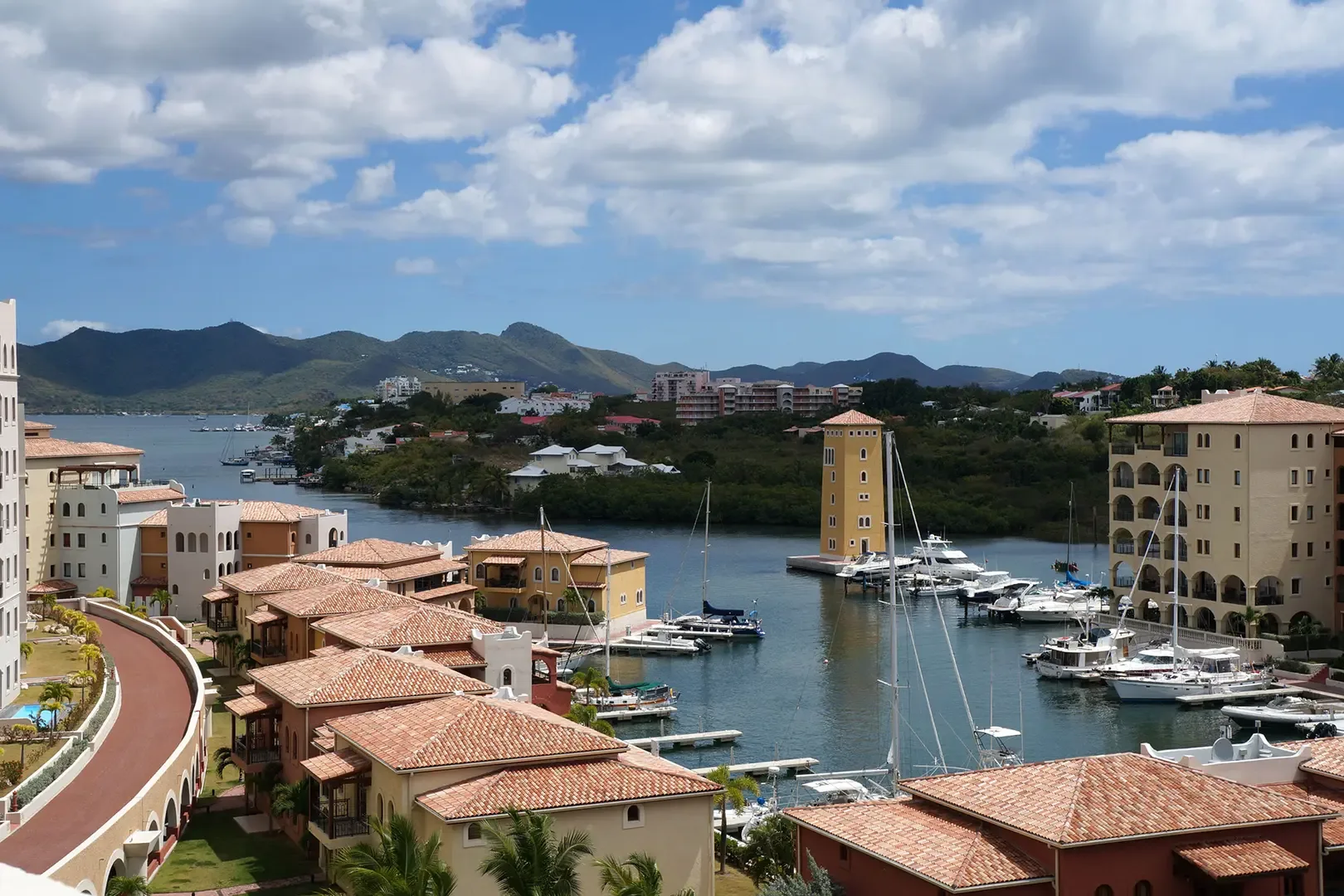 A high-angle scenic view showcases the Mediterranean-style resort and marina with terra cotta roofs, yellow tower, numerous yachts, mountains and blue sky backdrop.