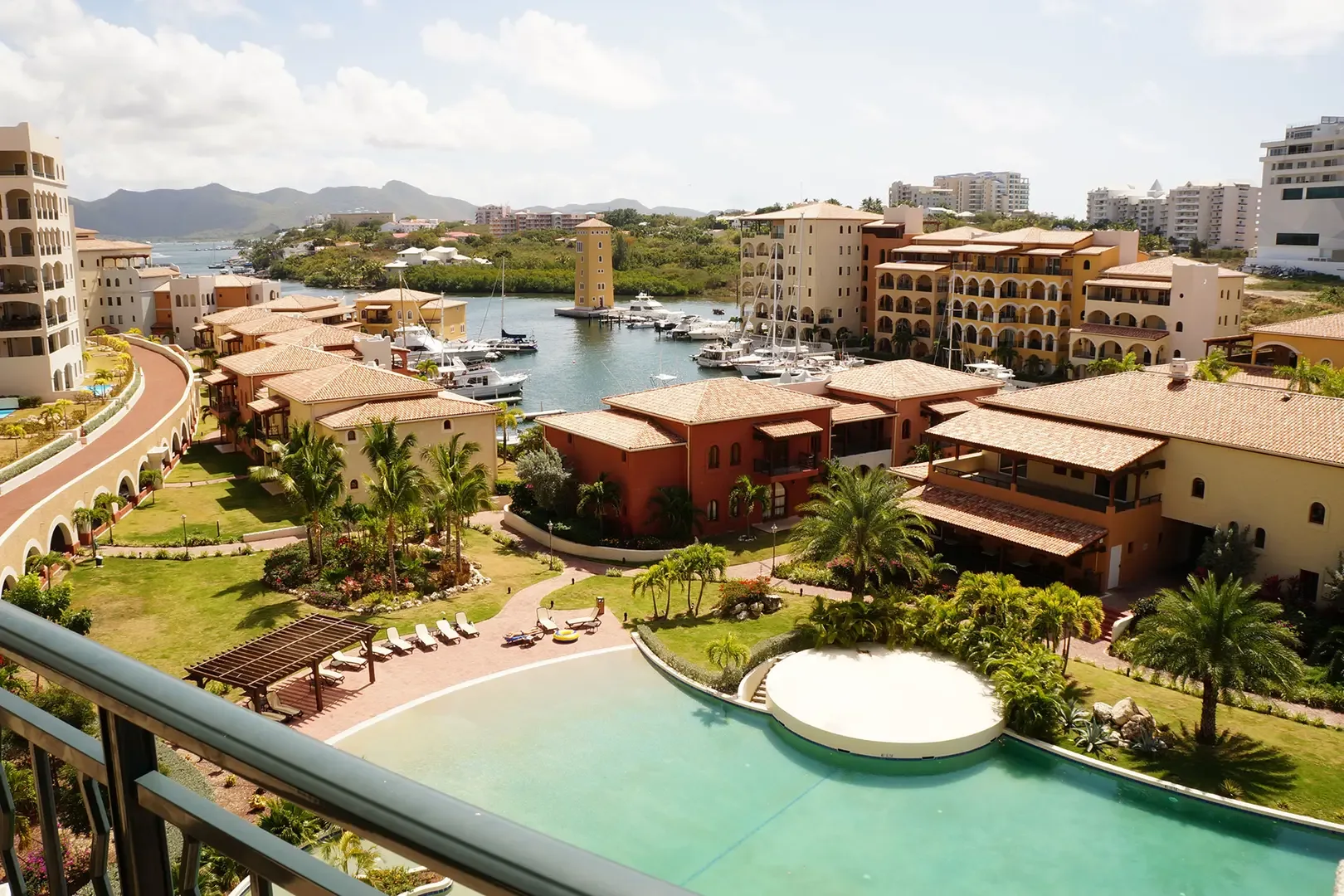 From your high-angle balcony, overlook the luxury resort complex with swimming pool, manicured gardens, marina with yachts, and Mediterranean architecture against mountains and bay.