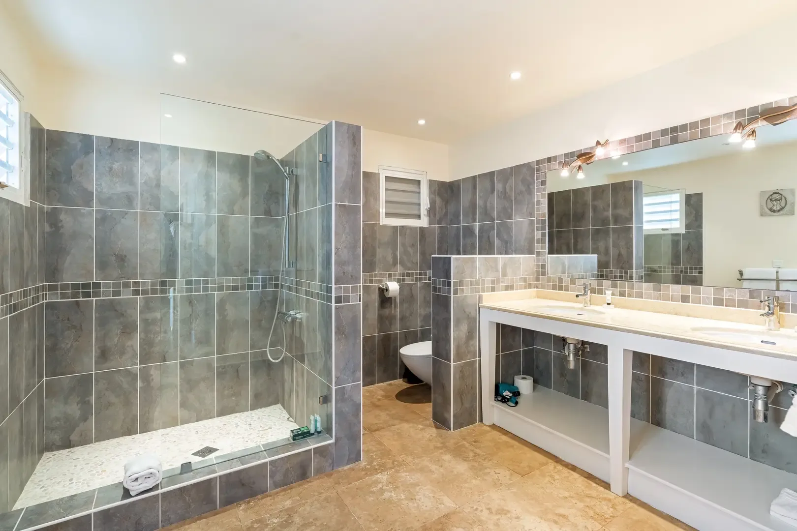 A spacious modern bathroom with walk-in pebble-floor shower, double vanity, and private toilet nook — spa-style relaxation at home.