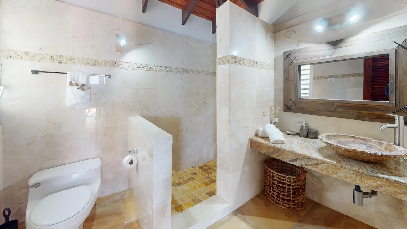 Modern bathroom with beige marble walls, rustic stone vessel sink, wooden-framed mirror, and gold-toned mosaic tile shower area