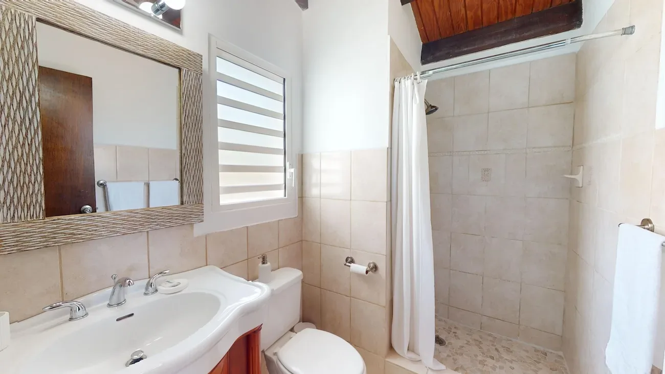 Bright bathroom with white sink, woven-frame mirror, shower with white curtain, and wooden ceiling detail