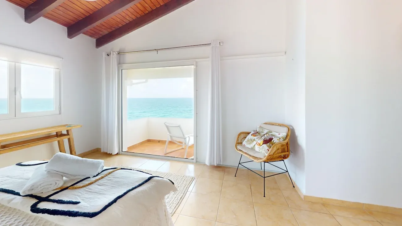 Bright bedroom with wooden ceiling beams, white linens with blue accents, wicker chair, and sliding glass doors to balcony with ocean view