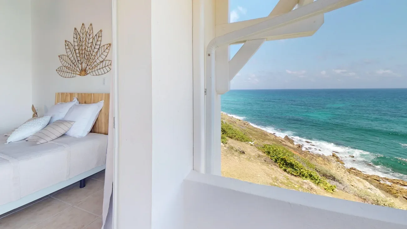 Bedroom with light bedding and wicker wall art, overlooking turquoise ocean and rocky coastline