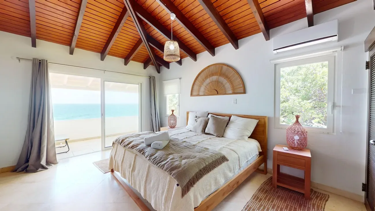Spacious bedroom with wooden ceiling, large bed, ocean view through sliding glass doors, and wicker decor