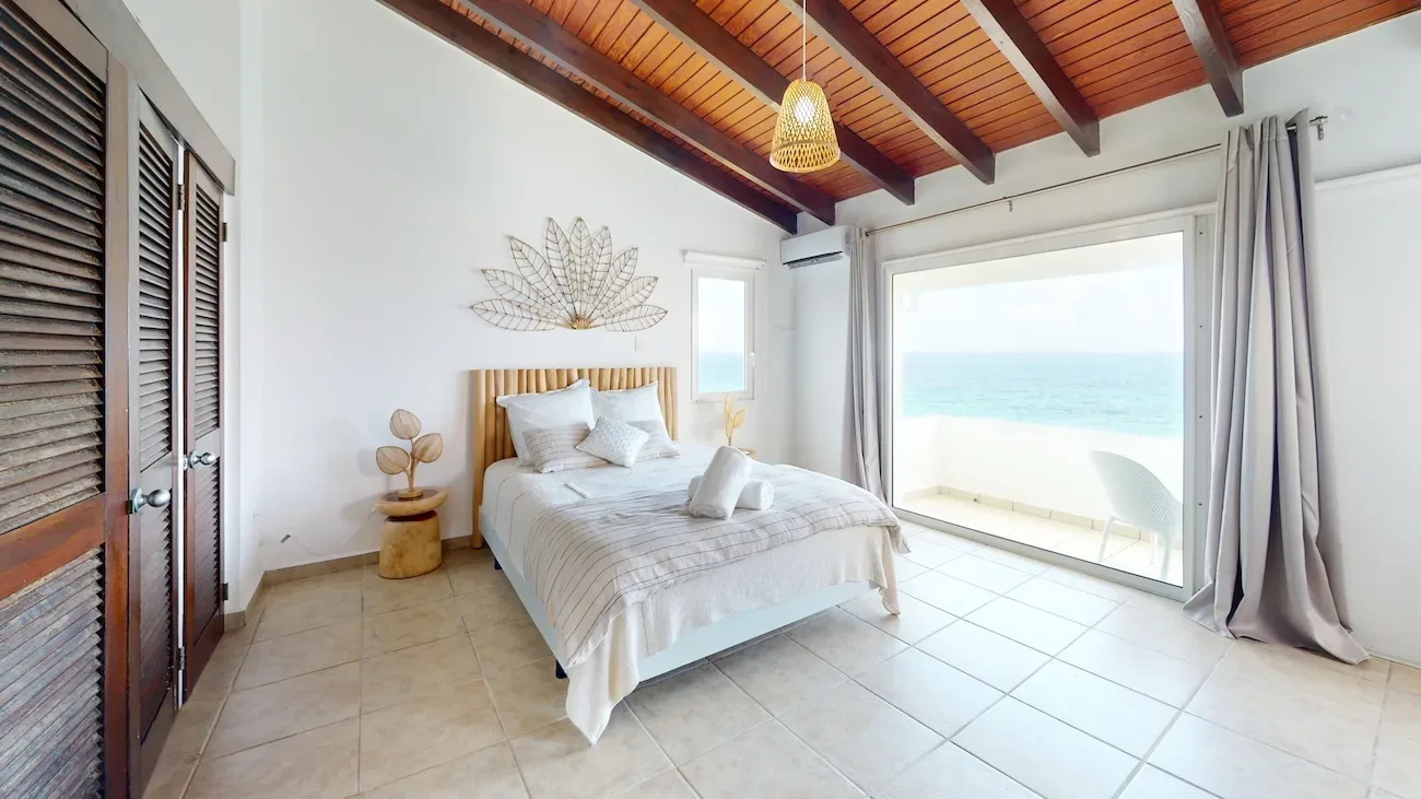 Spacious bedroom with ocean view through sliding glass doors, bed with striped linens, wooden ceiling beams, and tropical decor