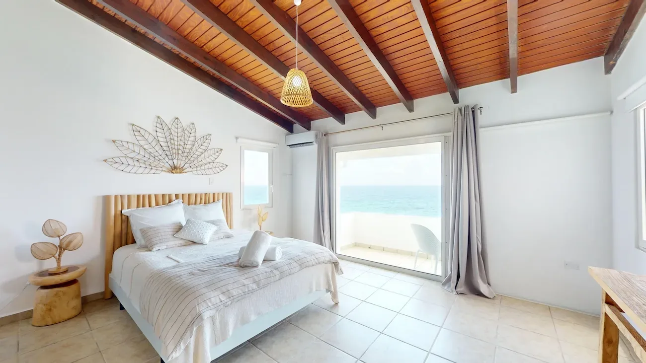 Spacious bedroom with ocean view, bamboo headboard, wooden ceiling beams, and light tile flooring