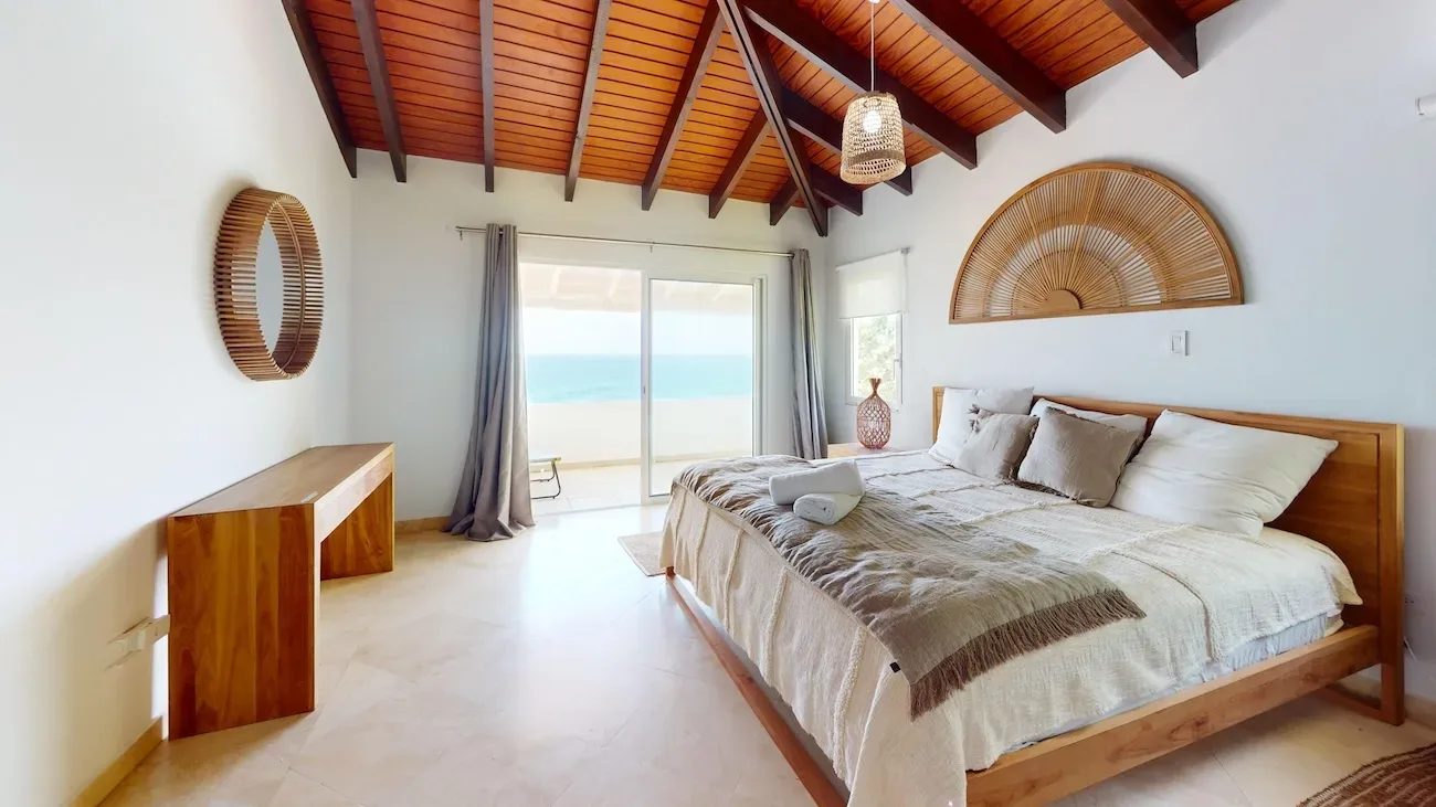 Spacious bedroom with wooden bed, ocean view through sliding glass doors, white walls, wooden ceiling beams, and coastal decor
