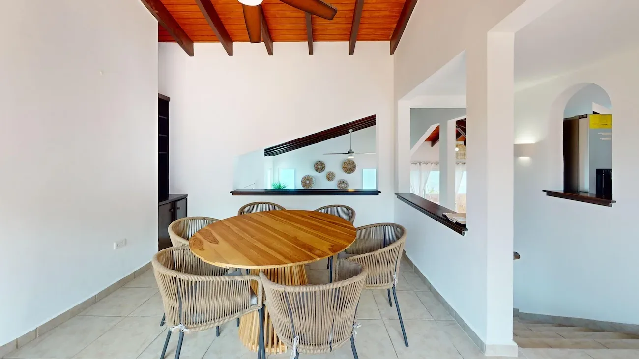 Bright dining area with round wooden table, six wicker chairs, white walls, exposed wooden ceiling beams, and ocean view