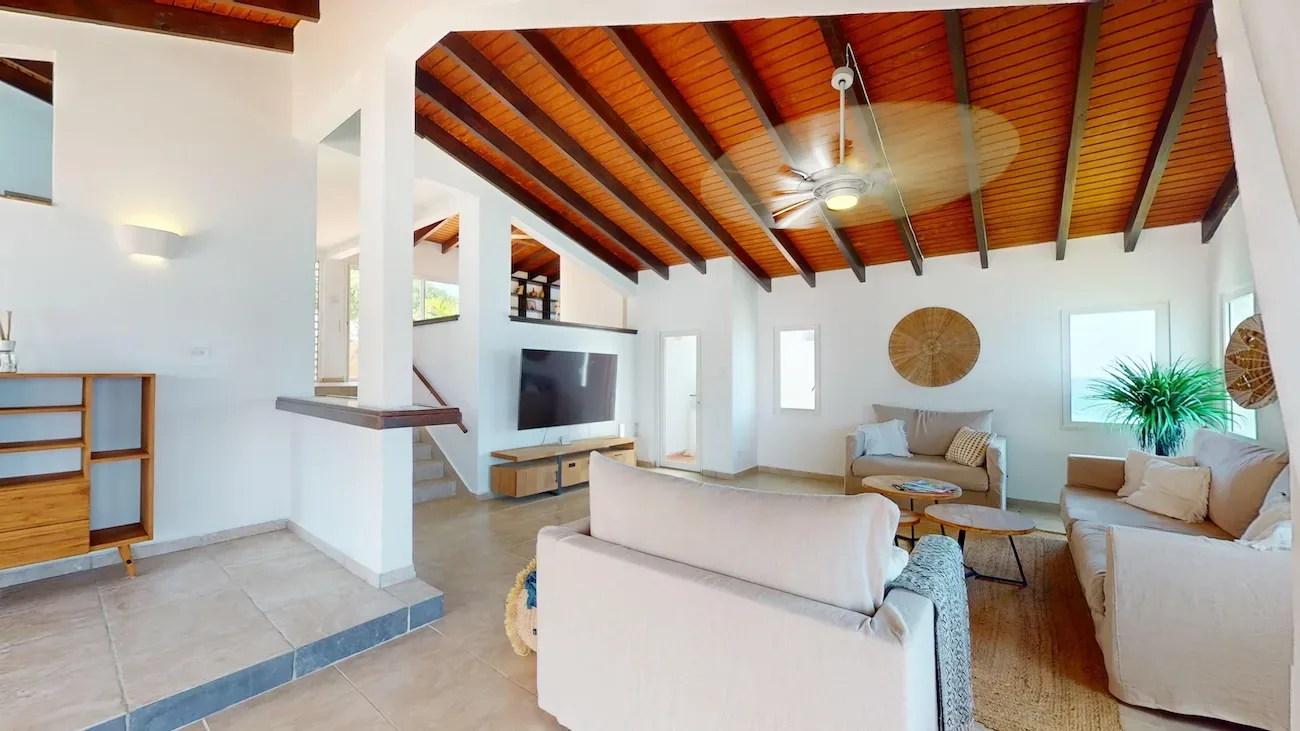Spacious living room with high wooden ceiling beams, beige sofas, flat-screen TV, round wooden coffee tables, and natural light