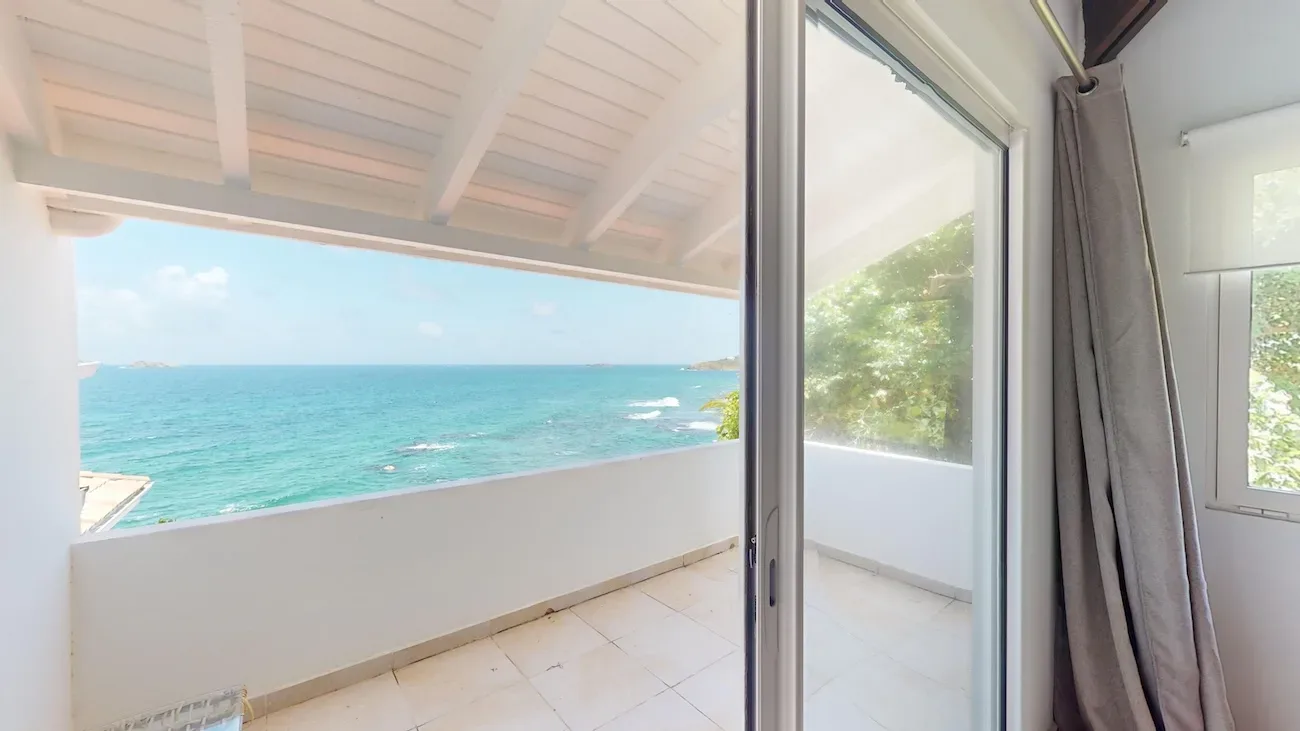 Bright indoor room with sliding glass doors opening to a covered balcony showcasing turquoise ocean waters