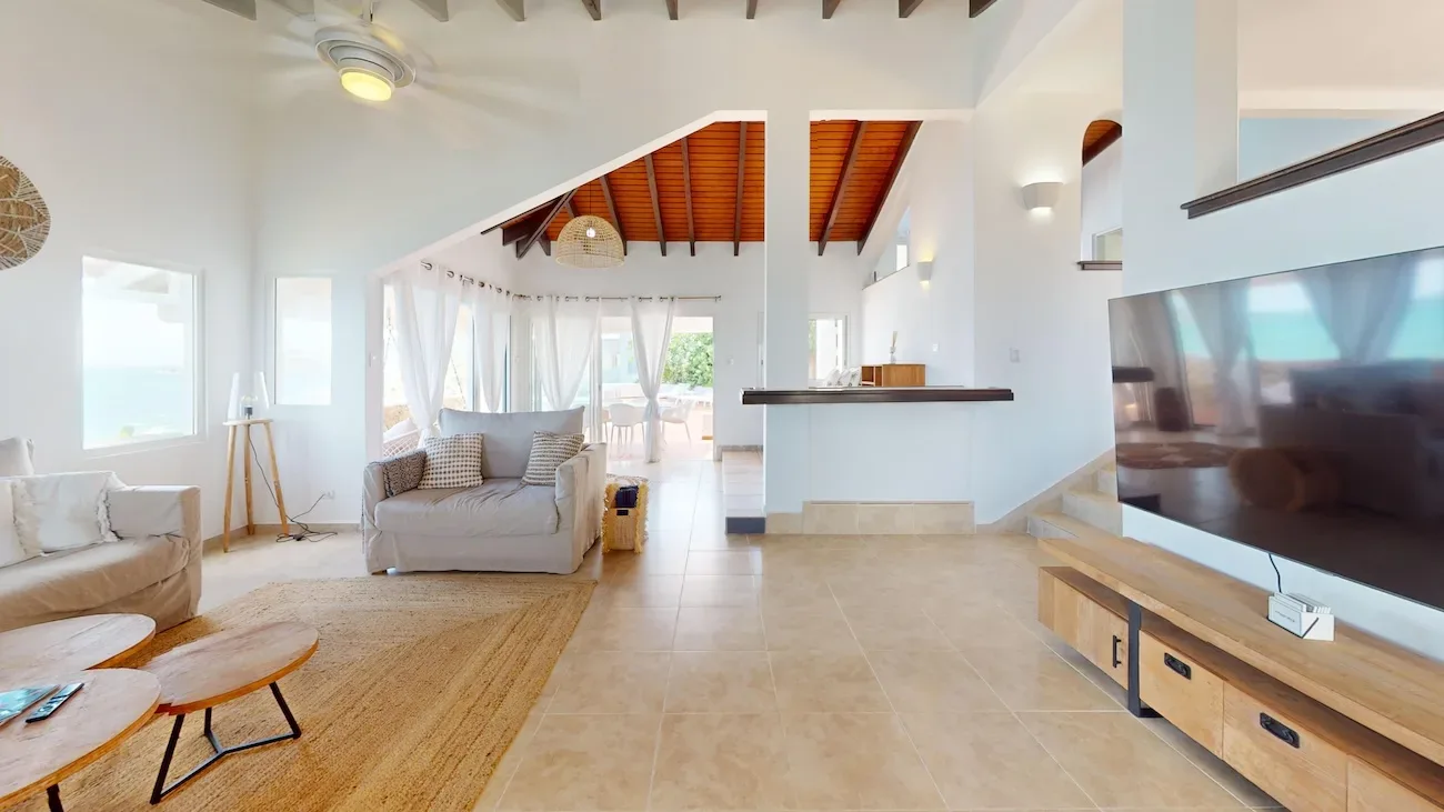 Spacious living room with ocean view, white walls, wooden ceiling beams, gray sofa, kitchen bar, and wooden coffee tables