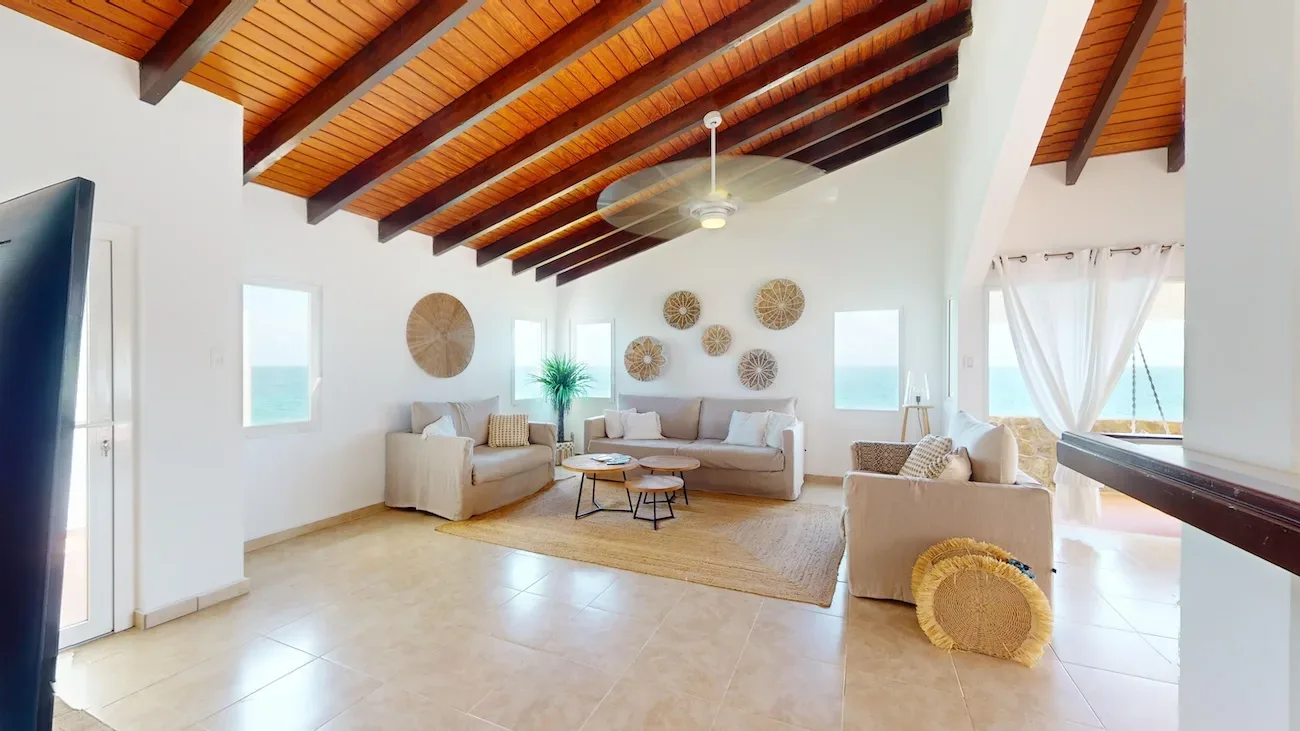 Spacious living room with beige sofas, wooden ceiling beams, ocean views, and coastal decor