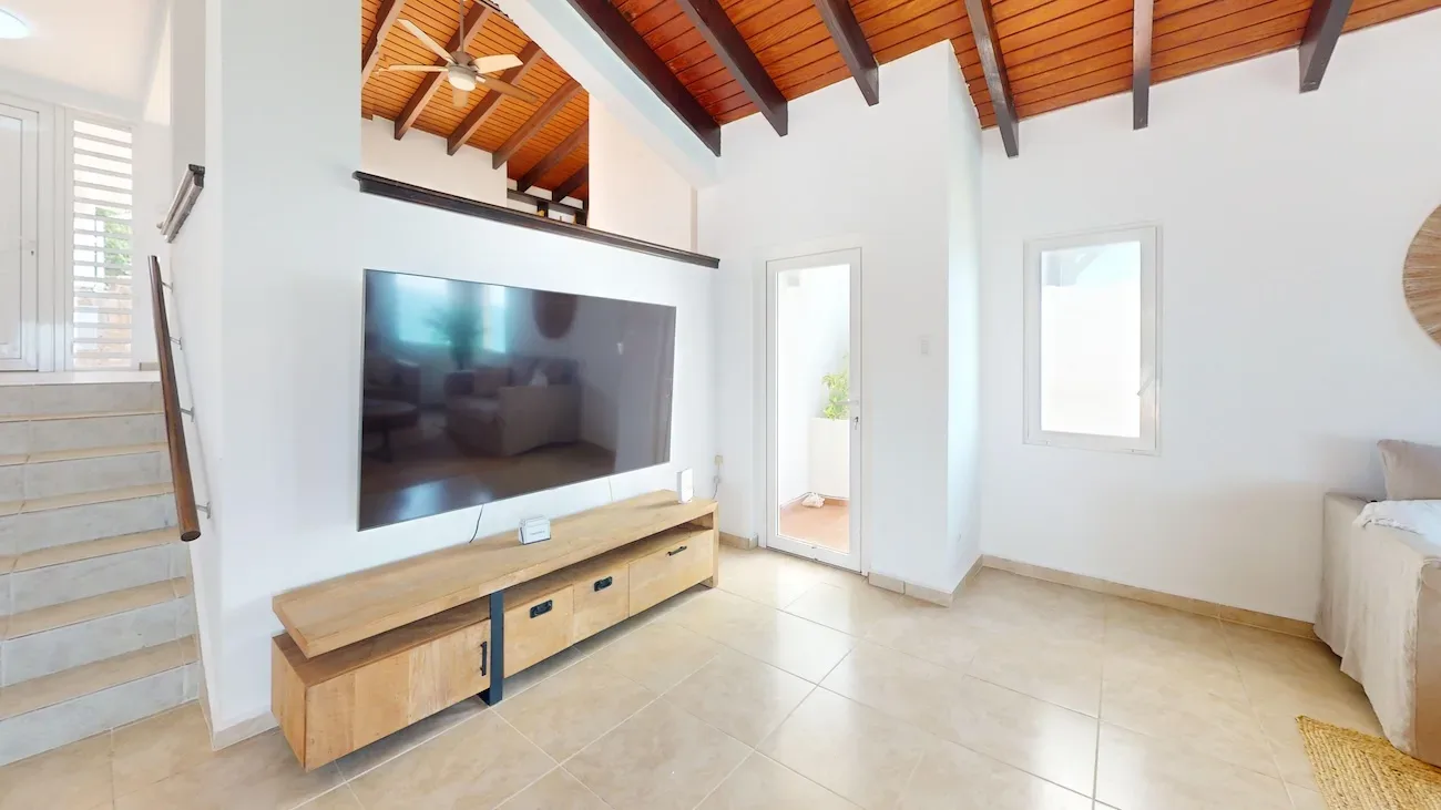 Bright living room with exposed wooden ceiling beams, large flat-screen TV, stone staircase, and access to outdoor area
