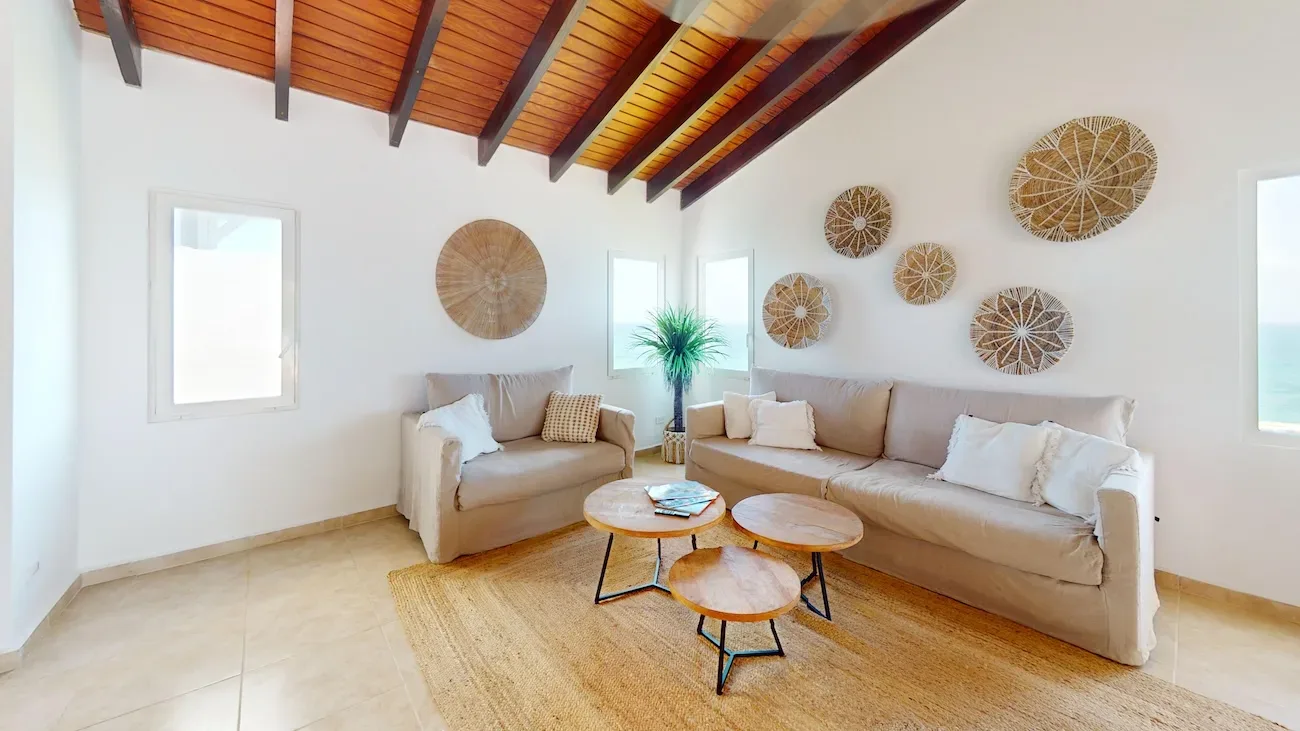 Spacious living room with beige sofas, woven wall art, wooden coffee tables, and ocean view through windows