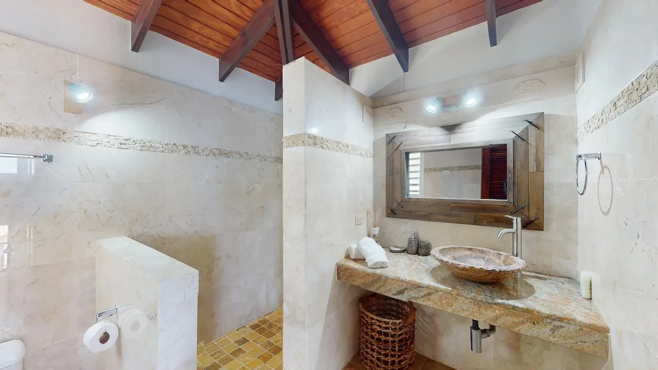 Luxury bathroom with beige marble walls, exposed wooden ceiling beams, stone vessel sink on granite countertop, and yellow mosaic shower floor