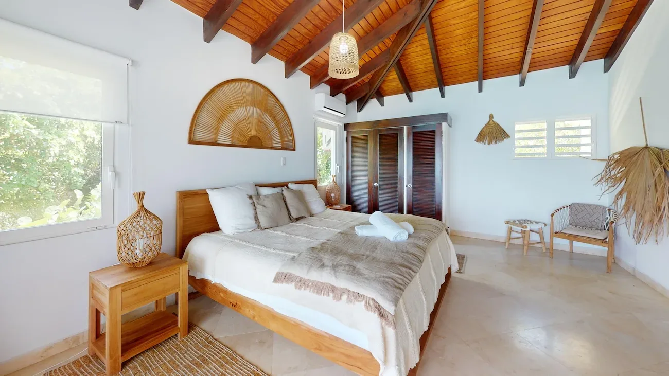 Spacious master bedroom with wooden ceiling beams, large bed, natural light from windows, and tropical decor