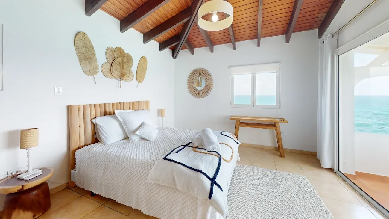 Ocean-view bedroom with large bed, wooden ceiling beams, palm leaf wall art, and sliding glass doors to the sea