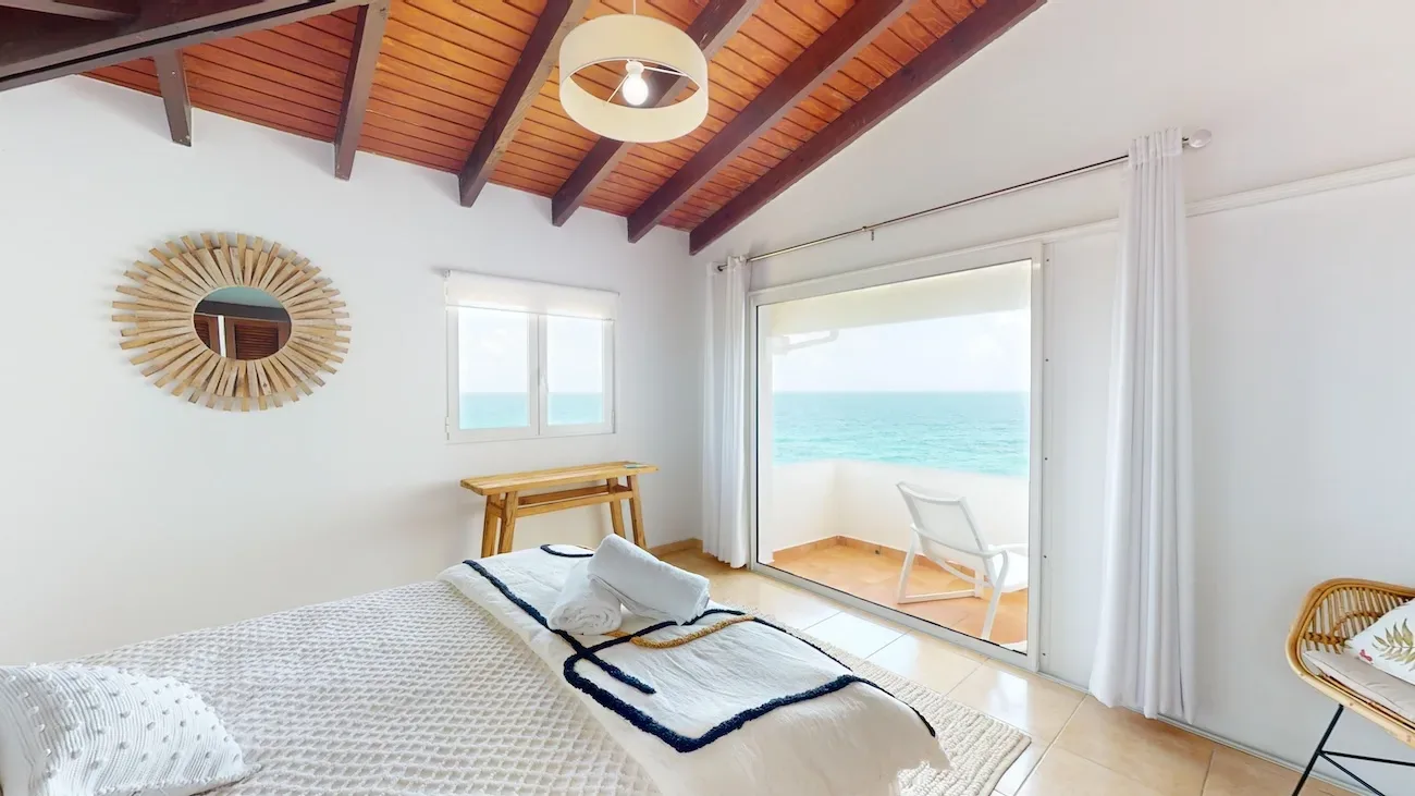 Bright bedroom with wooden ceiling beams and sliding doors opening to a balcony with turquoise ocean views
