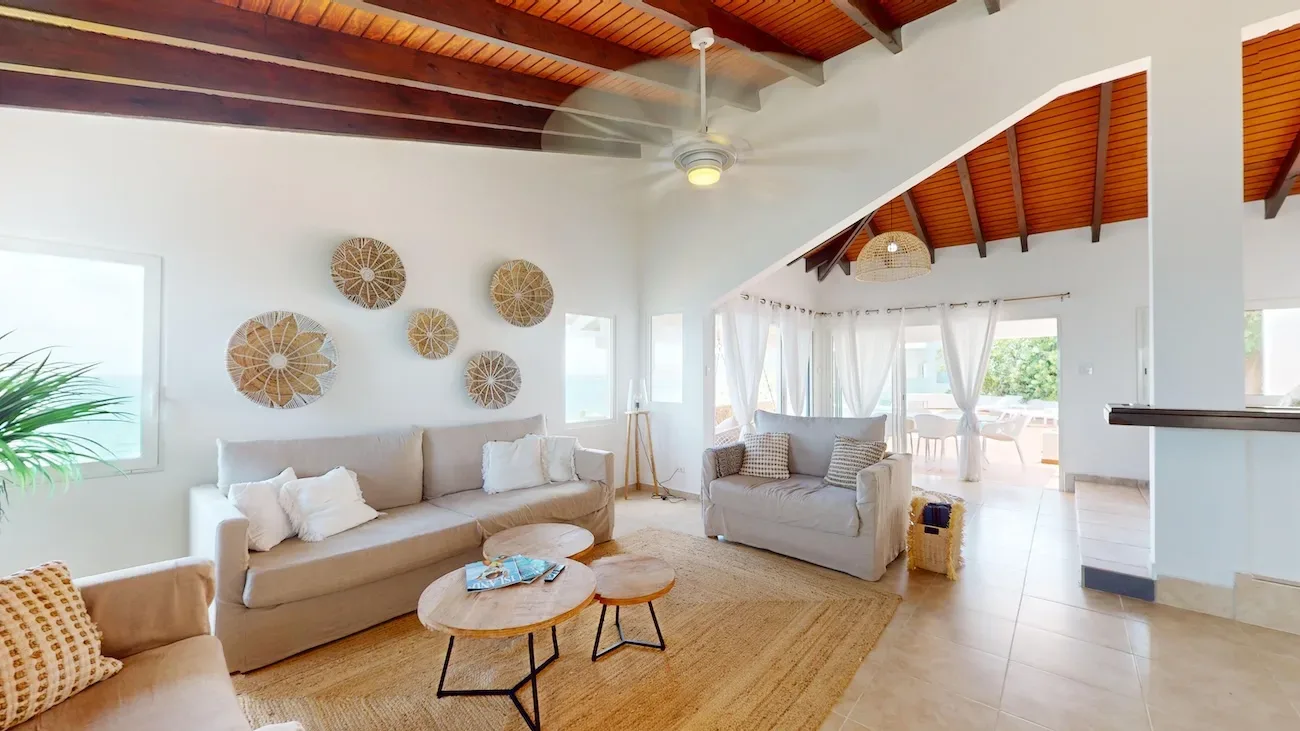 Spacious living room with beige sofas, wooden ceiling beams, woven wall art, and ocean view through large windows