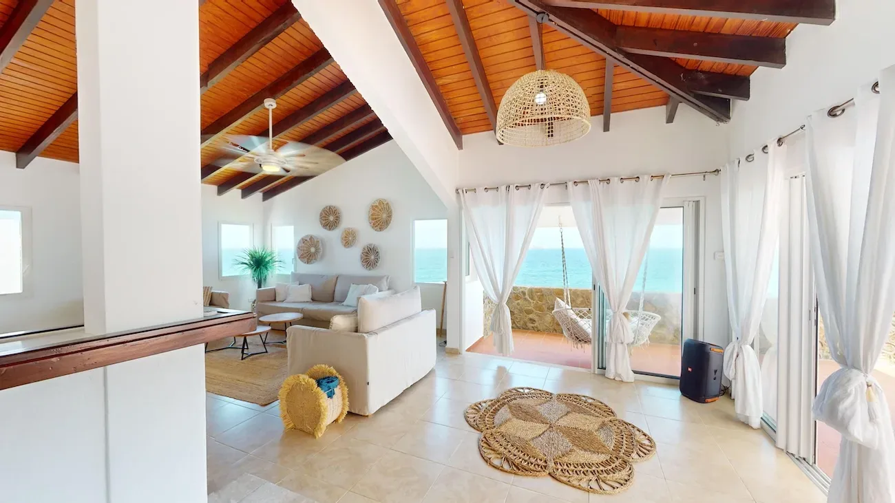 Bright open-concept living room with white walls, wooden ceiling beams, beige sofa, and ocean view through glass doors with hanging chairs