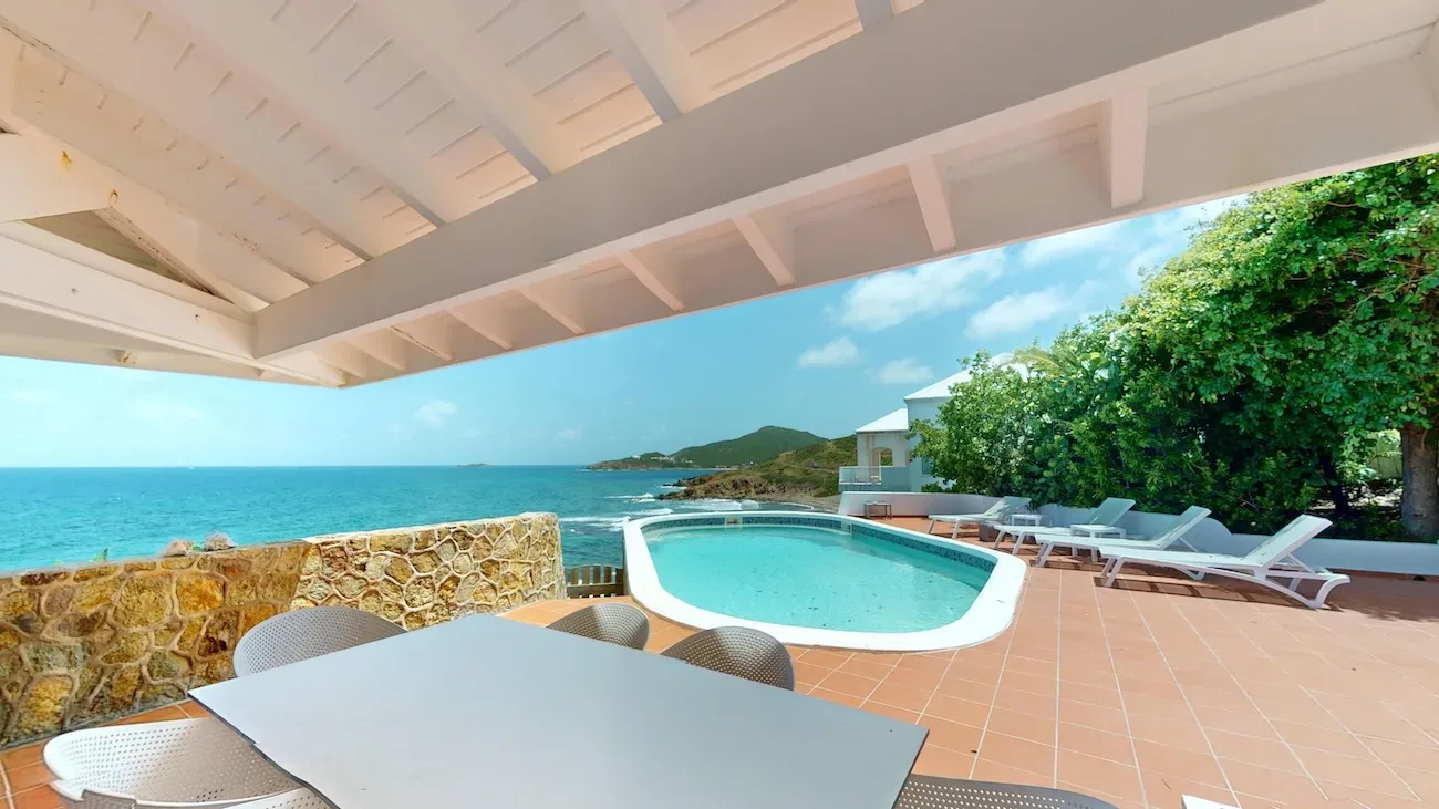 Covered terrace with white dining table, chairs, turquoise pool, and ocean view with stone wall and lush greenery