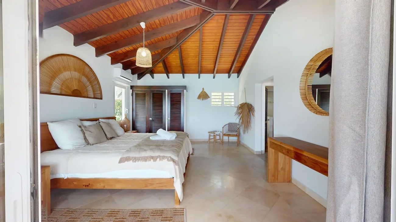 Spacious bedroom with wooden ceiling beams, large bed with beige linens, natural light, and tropical decor