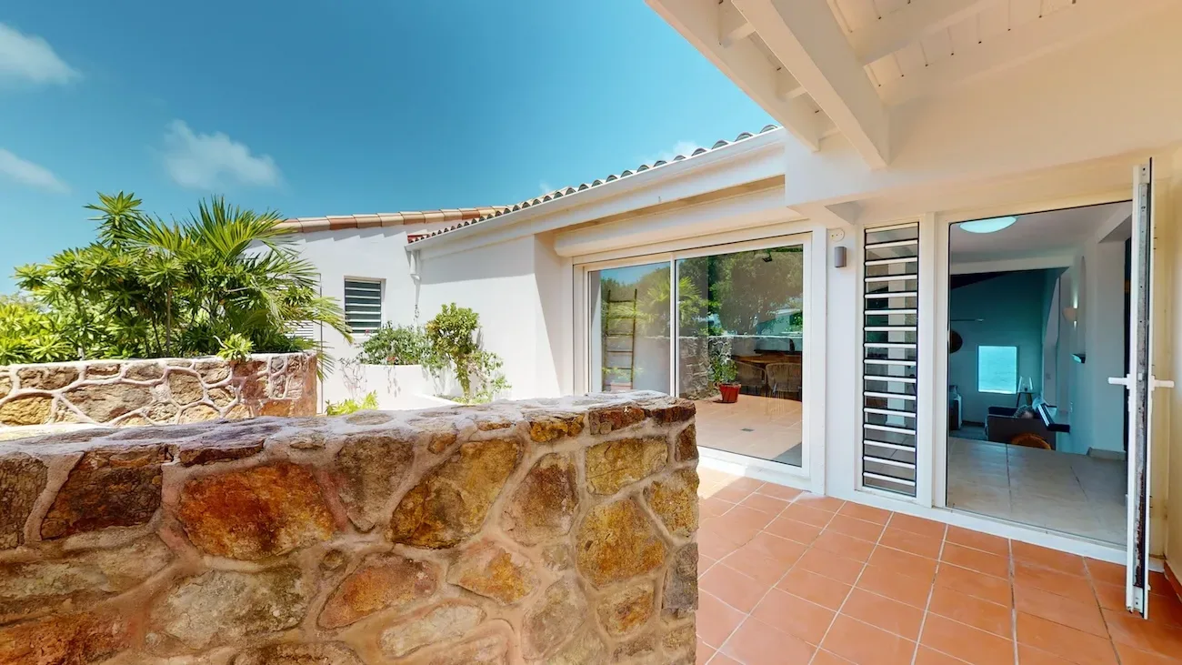 Villa outdoor patio with stone wall, terracotta tiles, sliding glass doors, tropical greenery, and clear blue sky