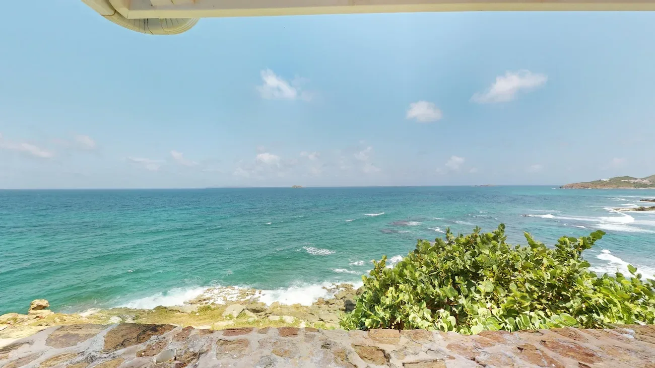 Ocean view from villa terrace with turquoise Caribbean Sea, rocky shoreline, green shrubs, and clear blue sky