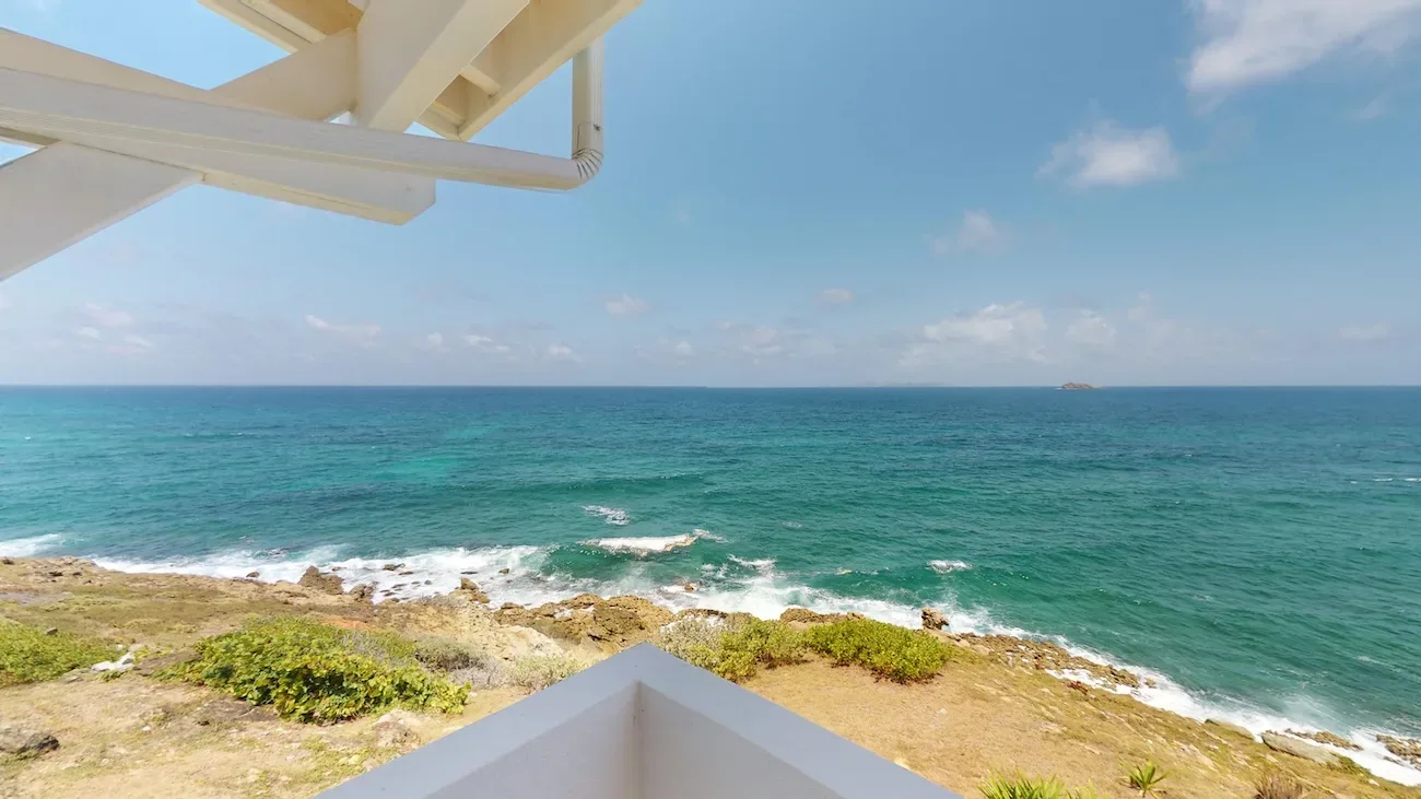 Villa terrace with white overhang overlooking turquoise ocean, rocky coastline, and clear blue sky