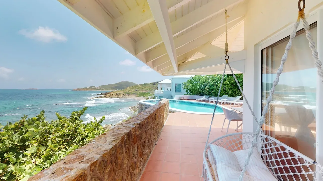 Terrace with white rope hammock, stone wall, pool, and ocean view at Timeless Tides villa