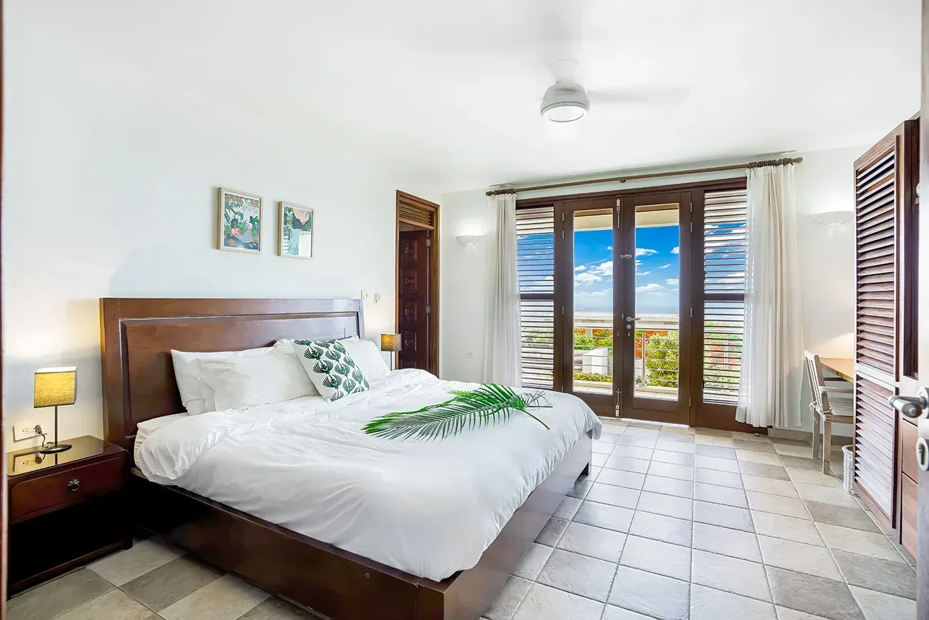 Spacious bedroom with king bed, white linens, wooden furniture, and ocean view through French doors