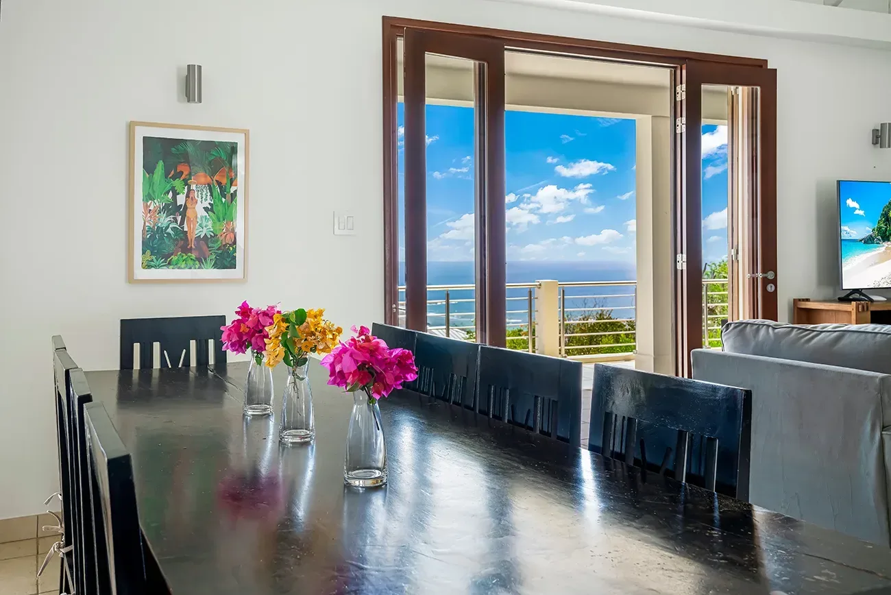 Dining area with dark table, colorful flowers in vases, open doors leading to ocean view, and living room beyond