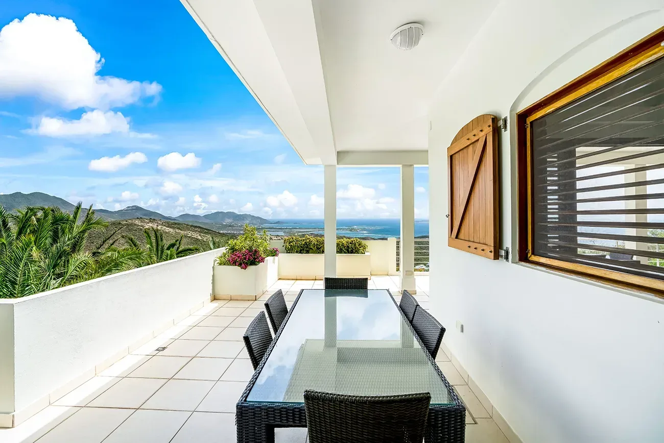 Glass-top dining table with wicker chairs on a covered terrace overlooking ocean and mountains
