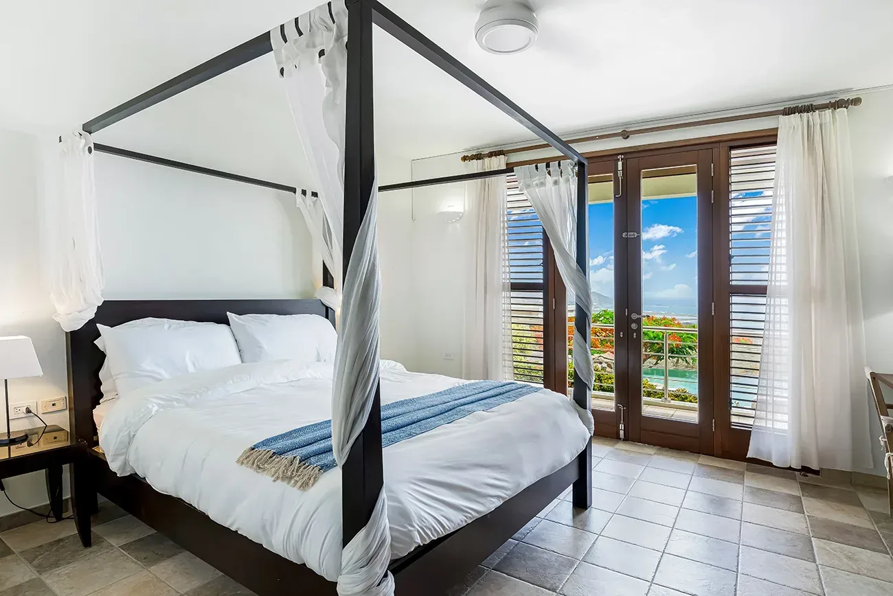Bedroom with four-poster bed, white linens, blue throw blanket, and panoramic ocean view through French doors