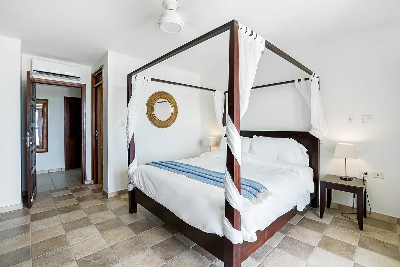 Bright bedroom with dark wooden four-poster bed, white linens, blue throw blanket, gold-framed mirror, and tiled flooring