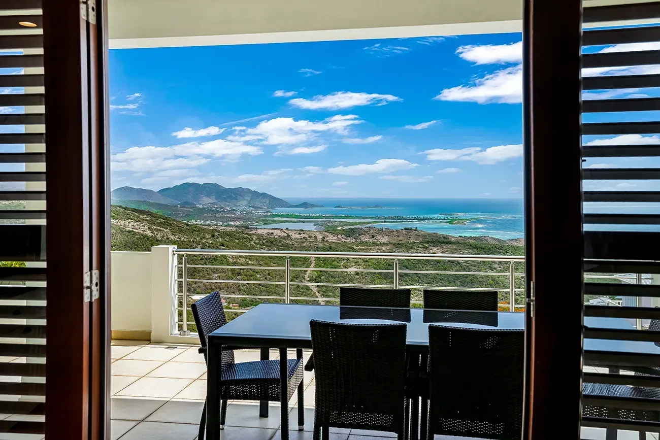 Villa interior with sliding doors opening to balcony with black wicker dining set and ocean views