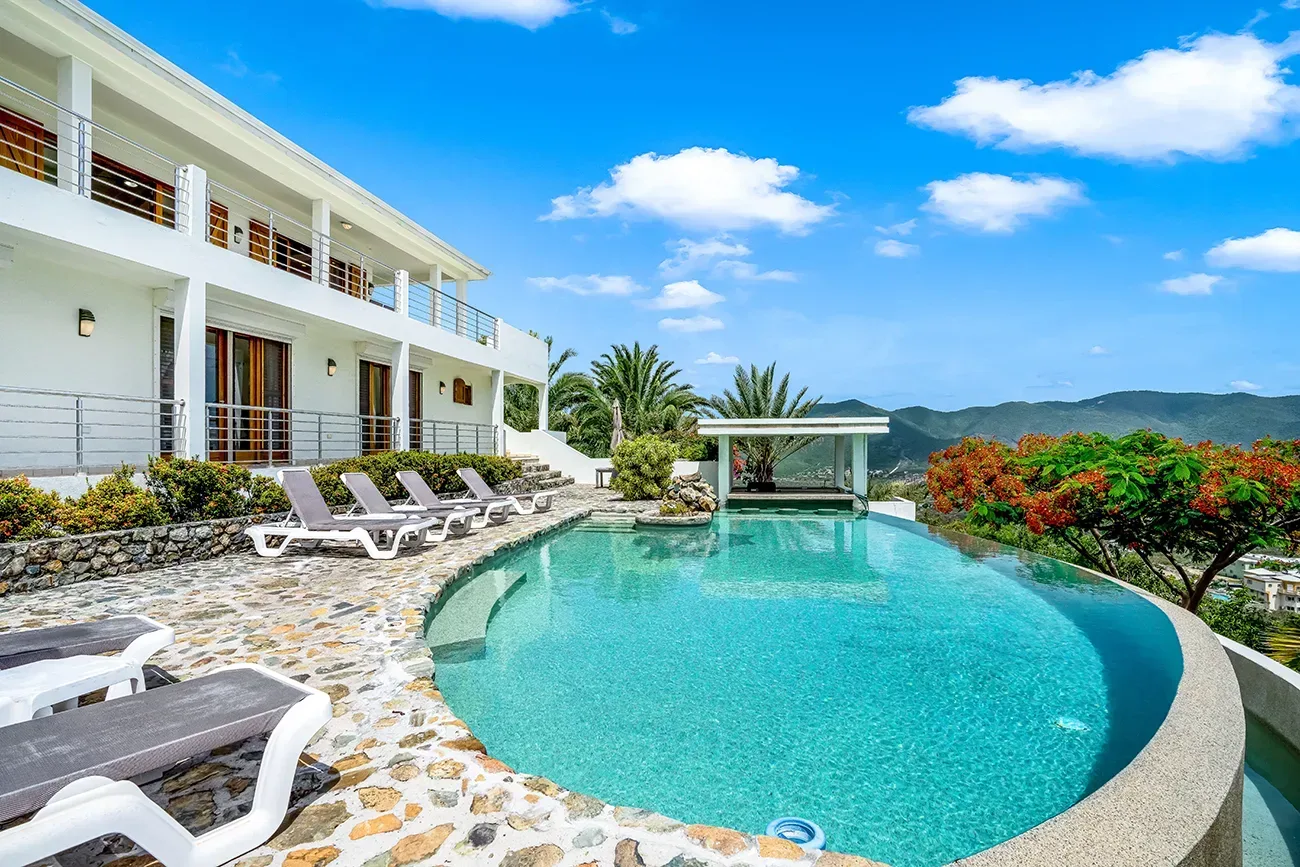 White two-story hillside villa with infinity pool, lounge chairs, stone patio, and panoramic mountain views