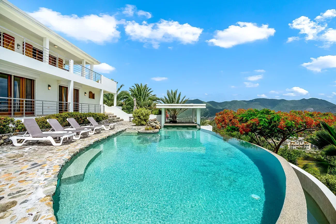 Modern villa with infinity pool, lounge chairs, mountain views, tropical plants, and clear blue sky