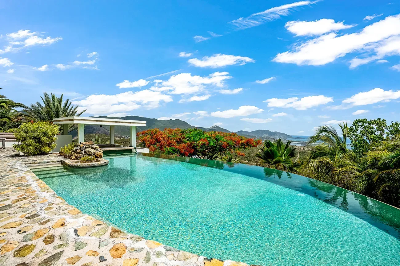 Luxury villa with turquoise infinity pool, stone deck, tropical landscaping, and mountain views under a bright blue sky